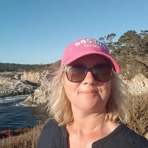 Jane Seale smiling outdoors, wearing dark sunglasses and a pink baseball cap, with the sea and rocky cliffs behind her.